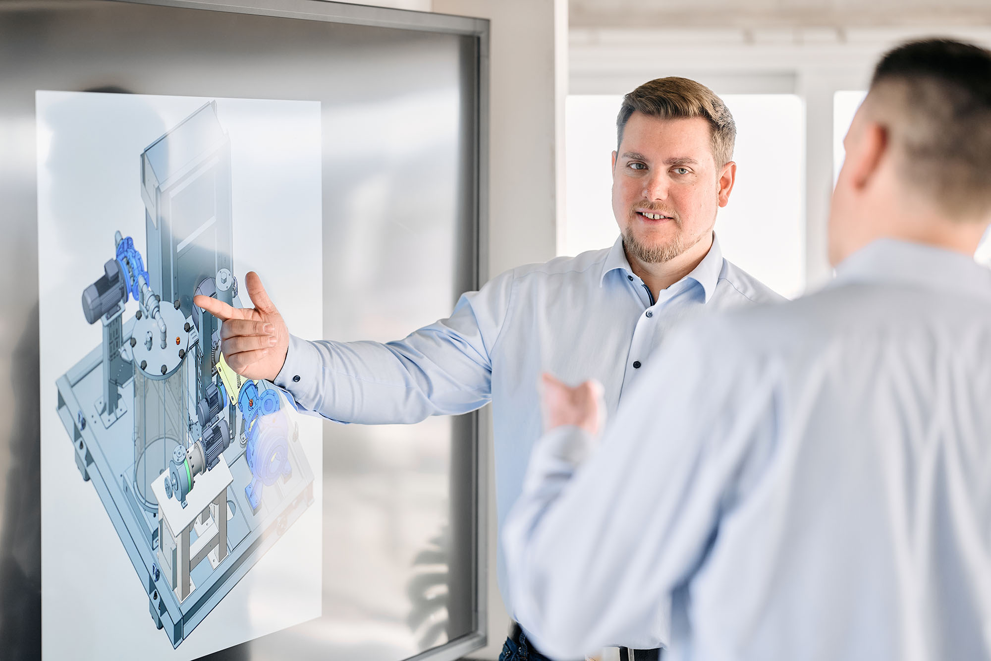 Mitarbeiter erklärt einem Kollegen eine technische Zeichnung auf einem Großbildschirm, Businessfotografie Köln