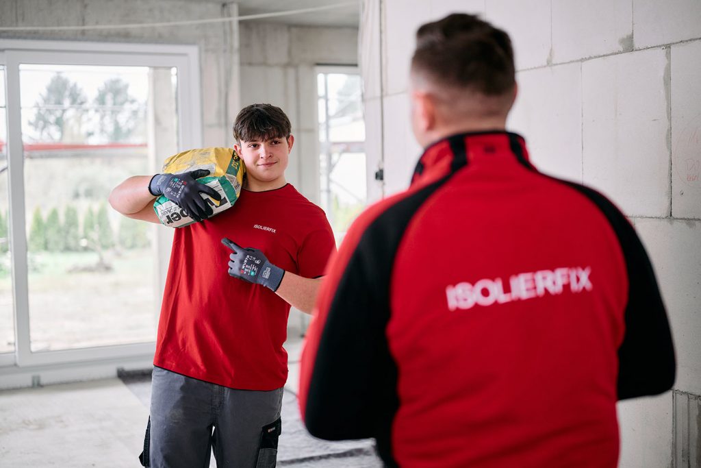 Ein Handwerker trägt einen Zementssack auf der Schulter und blickt lachend zu seinem Kollegen
