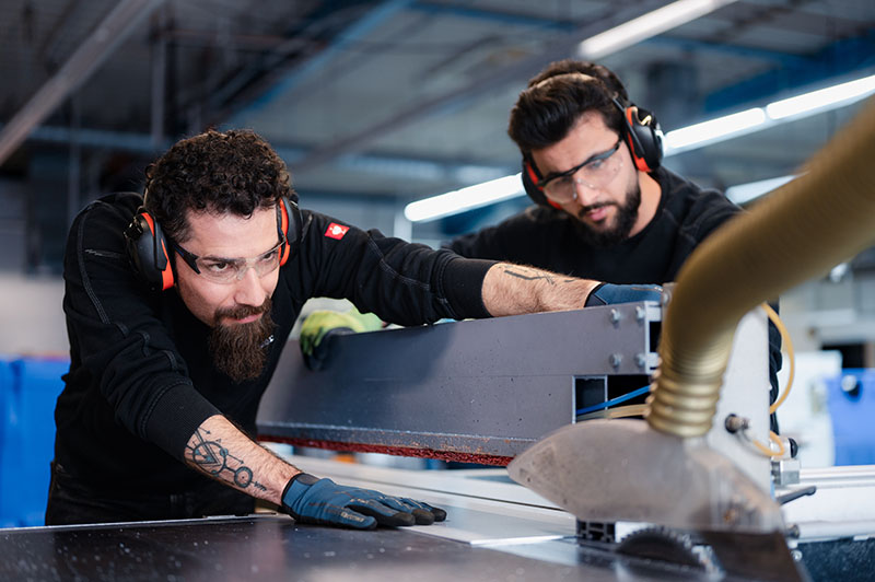 Zwei Industriearbeiter mit Gehörschutz und Schutzbrille arbeiten gemeinsam an einer Maschine in einer Kölner Produktionshalle