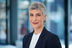 Modernes Mitarbeiterfoto einer Geschäftsfrau mit kurzem Haar im schwarzen Blazer vor Glasfassade